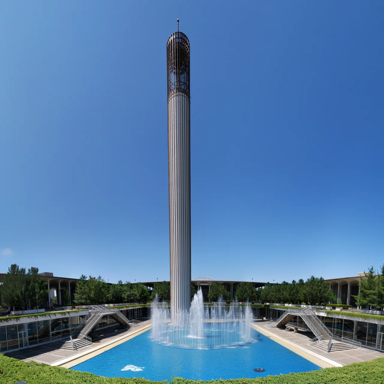 A tall fountain column rises above a blue pool, steps and colonnaded buildings at the University at Albany.