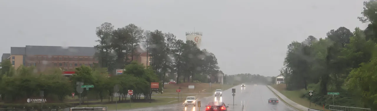 Rainy roadside view of Campbell University signs, campus buildings and the CU water tower in Buies Creek.