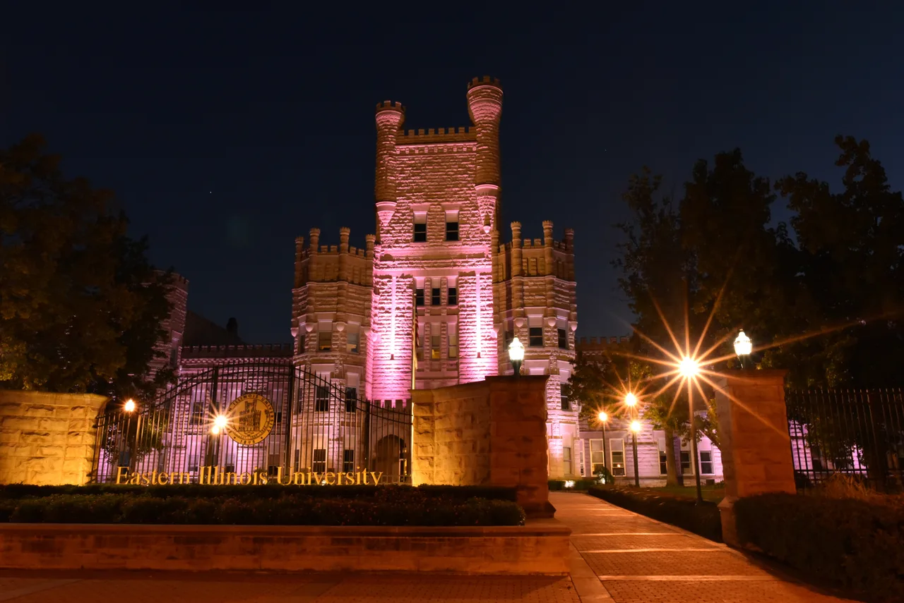 Illuminated Old Main tower above the Eastern Illinois University entrance sign and campus walkway at night.