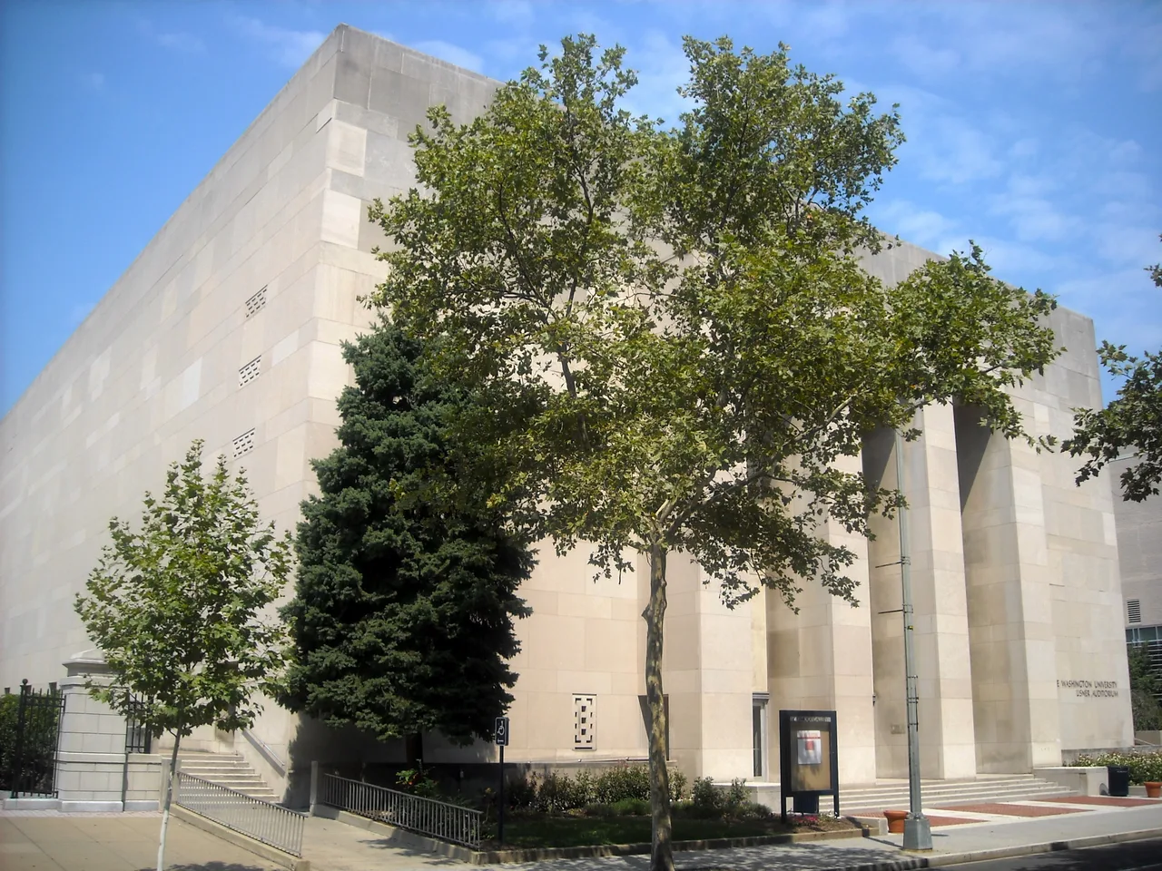 The pale stone exterior and entrance steps of George Washington University’s Lisner Auditorium, partly screened by a street tree.