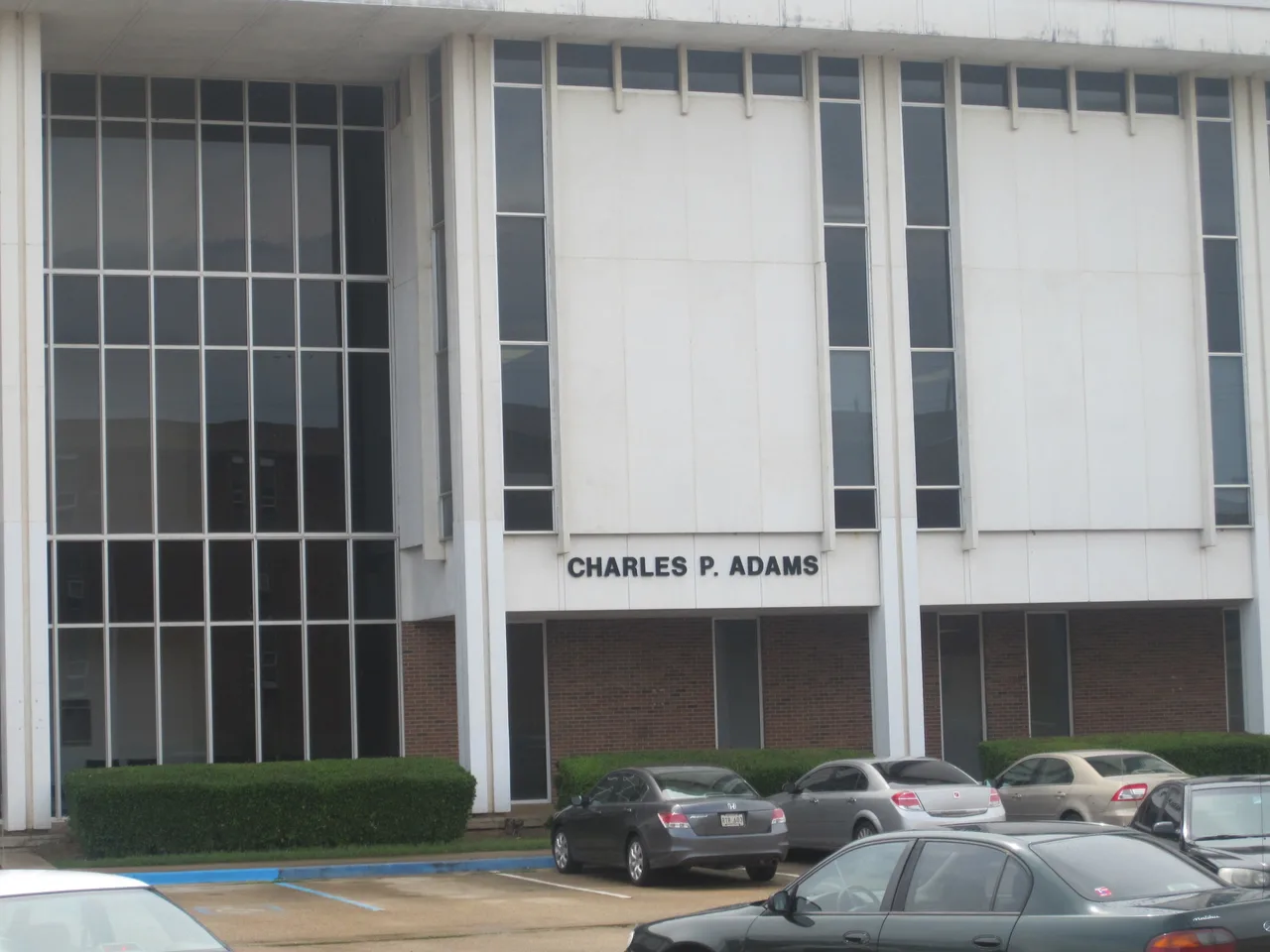 The white exterior panels and tall windows of Charles P. Adams Hall at Grambling State University, with its name above the entrance and parked cars below.