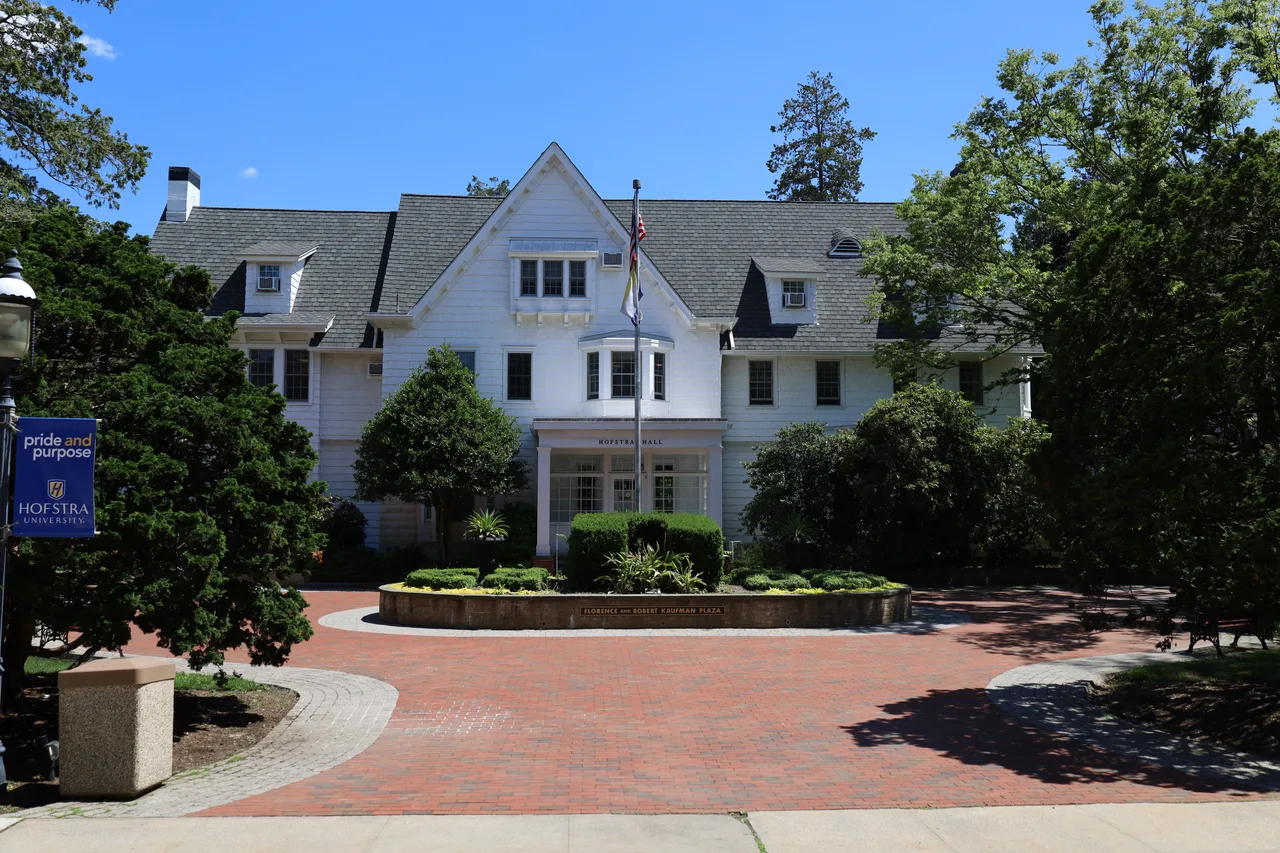 Hofstra Hall’s white gabled facade behind a landscaped brick plaza and flagpole at Hofstra University.