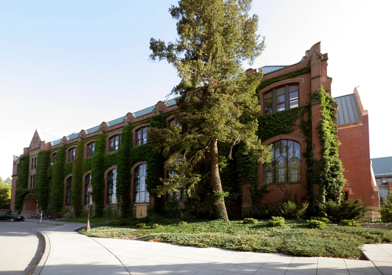 The ivy-covered brick exterior and arched windows of the University of Idaho Administration Building beside a campus walk, partly screened by an evergreen.
