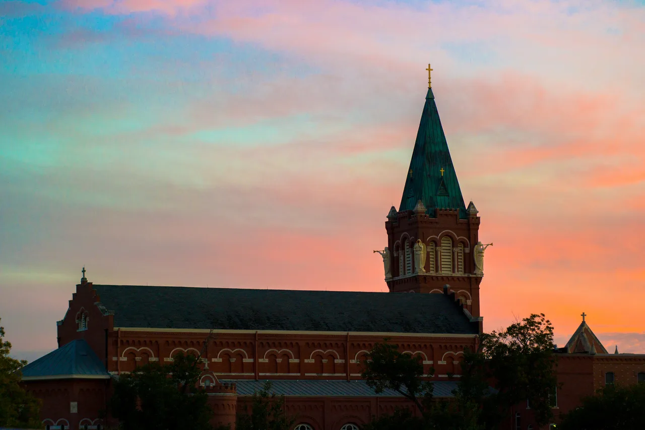 A brick campus building and cross-topped steeple beneath a colorful sunset at the University of the Incarnate Word in San Antonio.