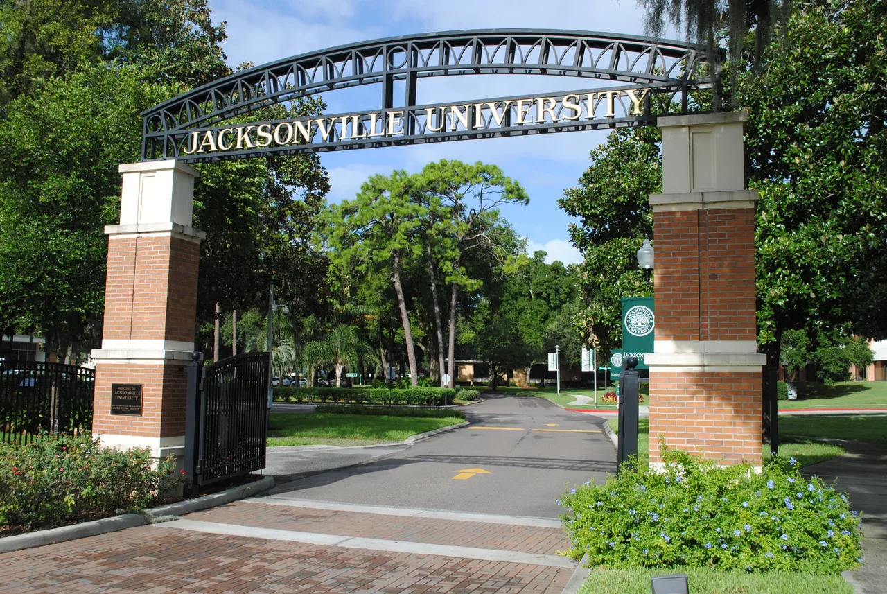 The Jacksonville University entrance arch above brick pillars, opening onto a tree-lined campus road in Jacksonville, Florida.